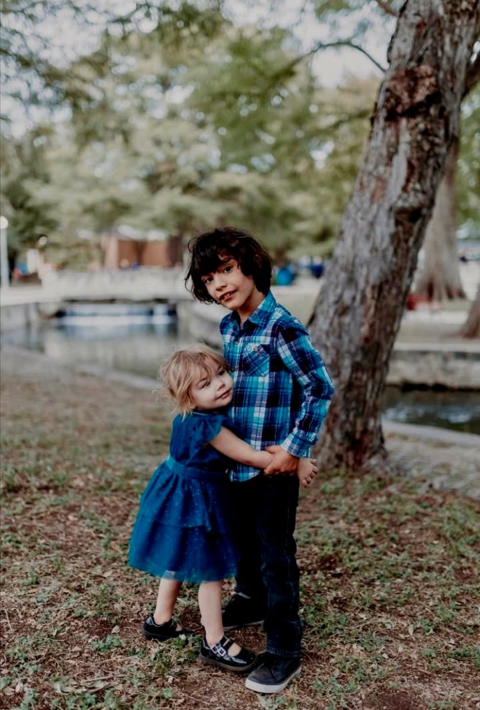 Brayden (on the right) stands with his sister, whose arms are wrapped around him in a hug. His sister wears a blue lacy dress, while Brayden wears a blue flannel shirt and nice blue plants. There is a tree and what looks like a canal in the background.