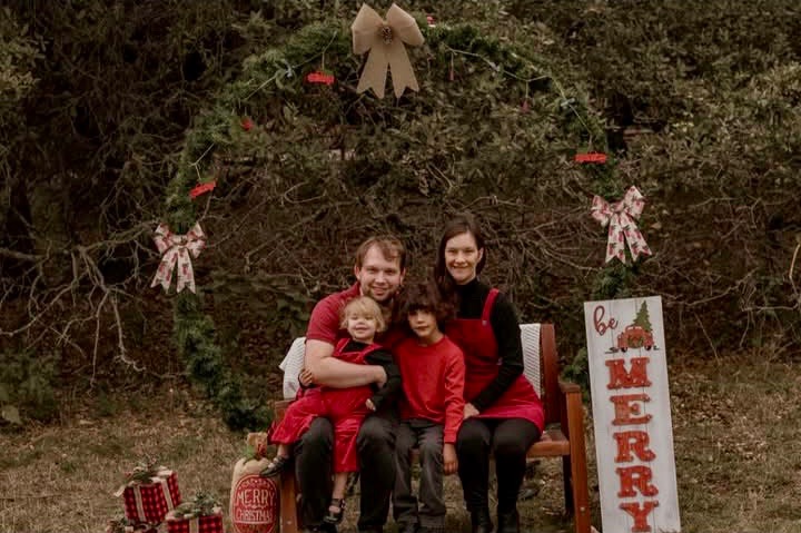 Hannah, her husband, and their two children sit in front of a wooded area. They are sitting on a brown bench framed by a large wreath. On the right side of the photo is a sign that reads: "Be Merry." On the left side of the photo there are several wrapped gifts. The family all wears red and smiles at the camera, excited about Christmas.
