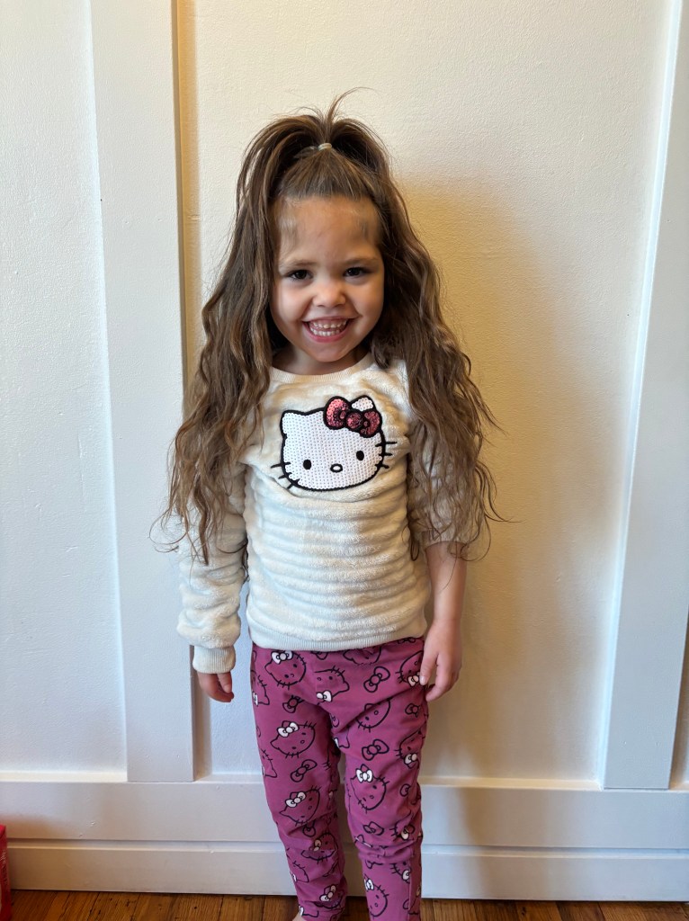 Malani, a young girl with BPAN, smiles at the camera. She stands in front of a white background. Her long brown hair cascades over her shoulders to her ellbows. She is wearing a white shirt with Hello Kitty on the front and pink pants with Hello Kitty.
