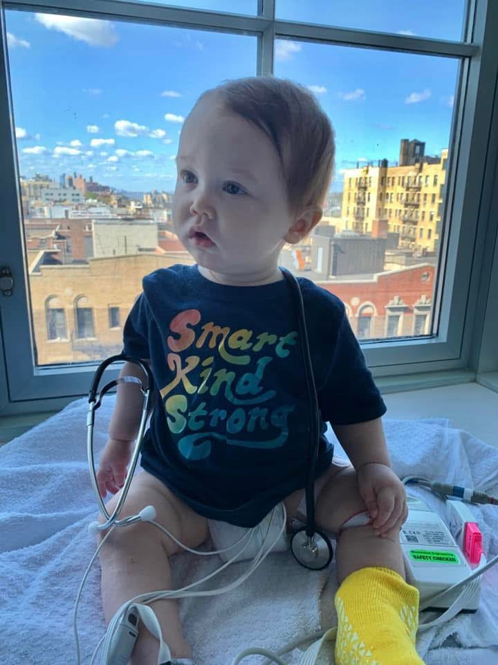 Noah sits at the doctor after his first cardiac arrest. He is wearing a black shirt that says "Smart, Kind, Strong" in rainbow letters. He has a stethoscope draped over his shoulders. 