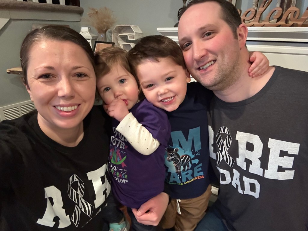 Kathleen, her daughter, her son Noah who has PPA2, and her husband all smile at the camera. Kathleen wears a shirt that reads: "Rare Mom." Her husband's shirt reads: "Rare Dad."
