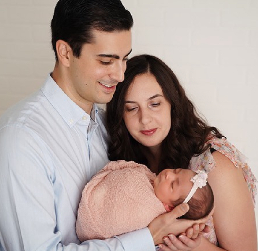 Frances and her husband John smile down at their daughter, Violet. Violet is a newborn swaddled in a pink blanket with a light pink headband.
